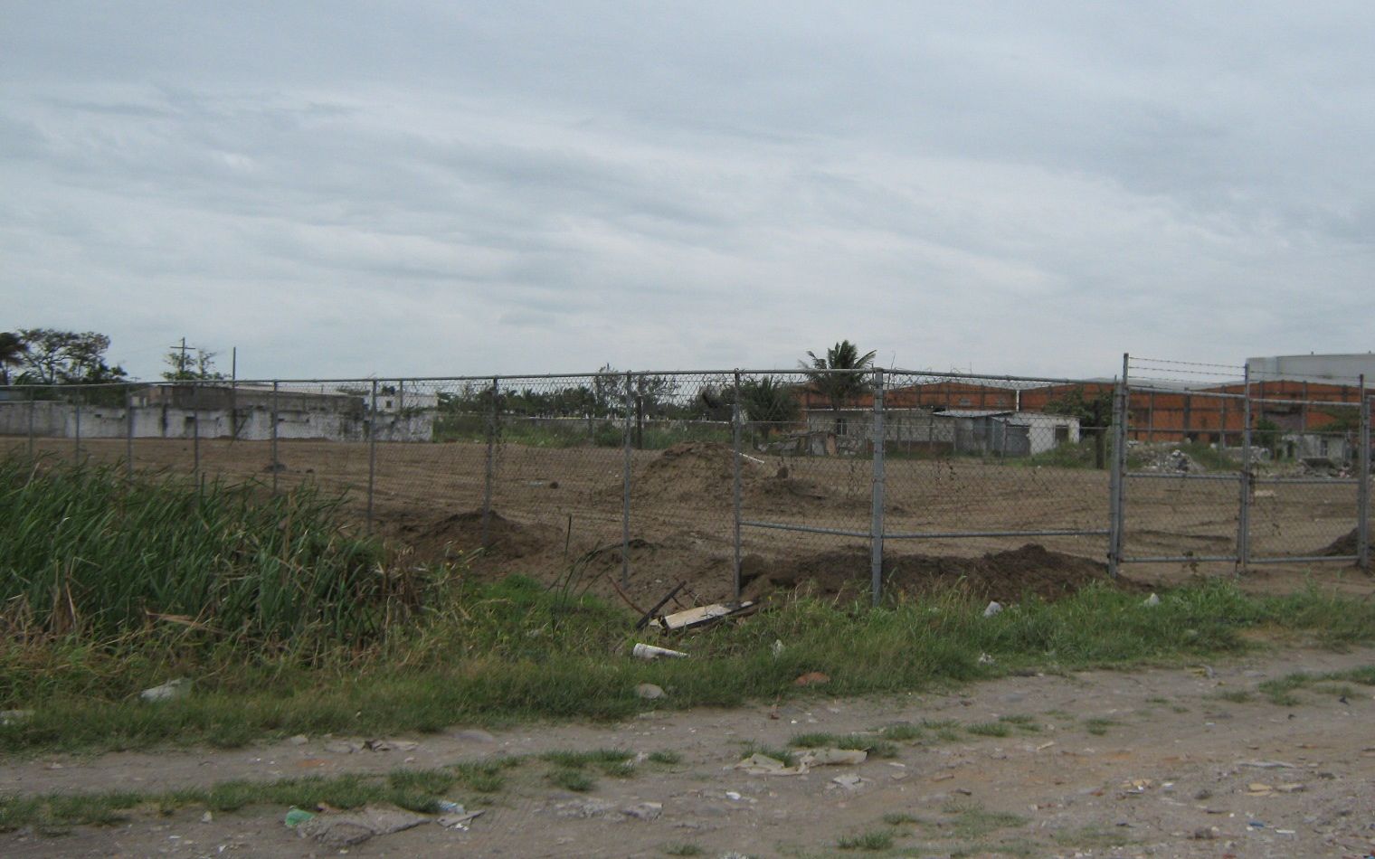 El terreno desde la calle en abril de 2009: plano, de tierra, con malla ciclónica y un portón de dos hojas, y bodegas y casas al fondo.
