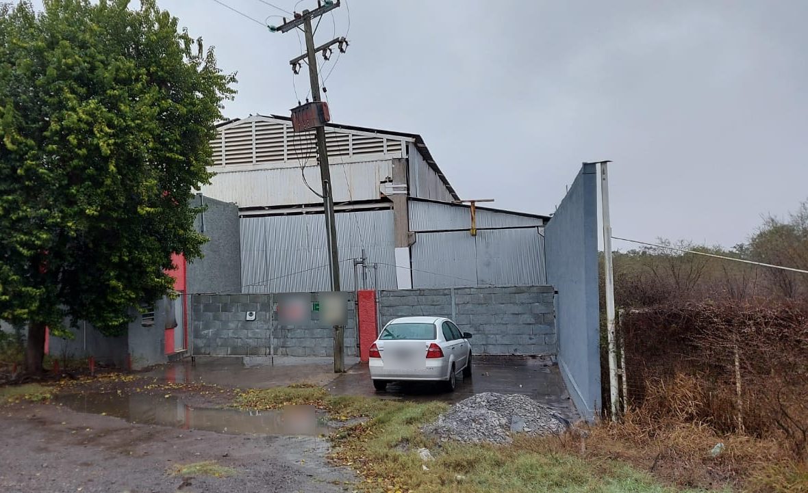 La planta vista desde la orilla de la carretera: barda de block al frente, poste de CFE, la nave detrás y un árbol grande junto al acceso.