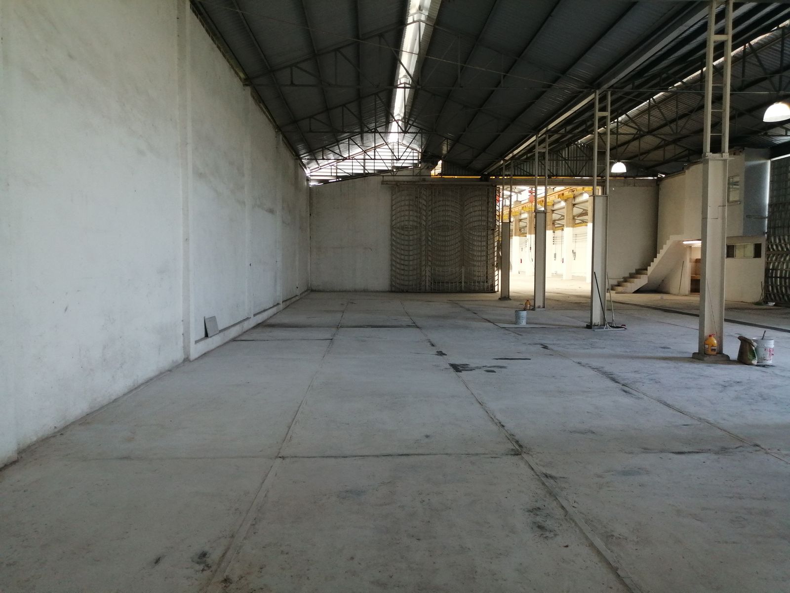 Nave posterior vacía, con muros de block pintados de blanco, columnas y armaduras de acero, firme de concreto con juntas y una reja de herrería al fondo.